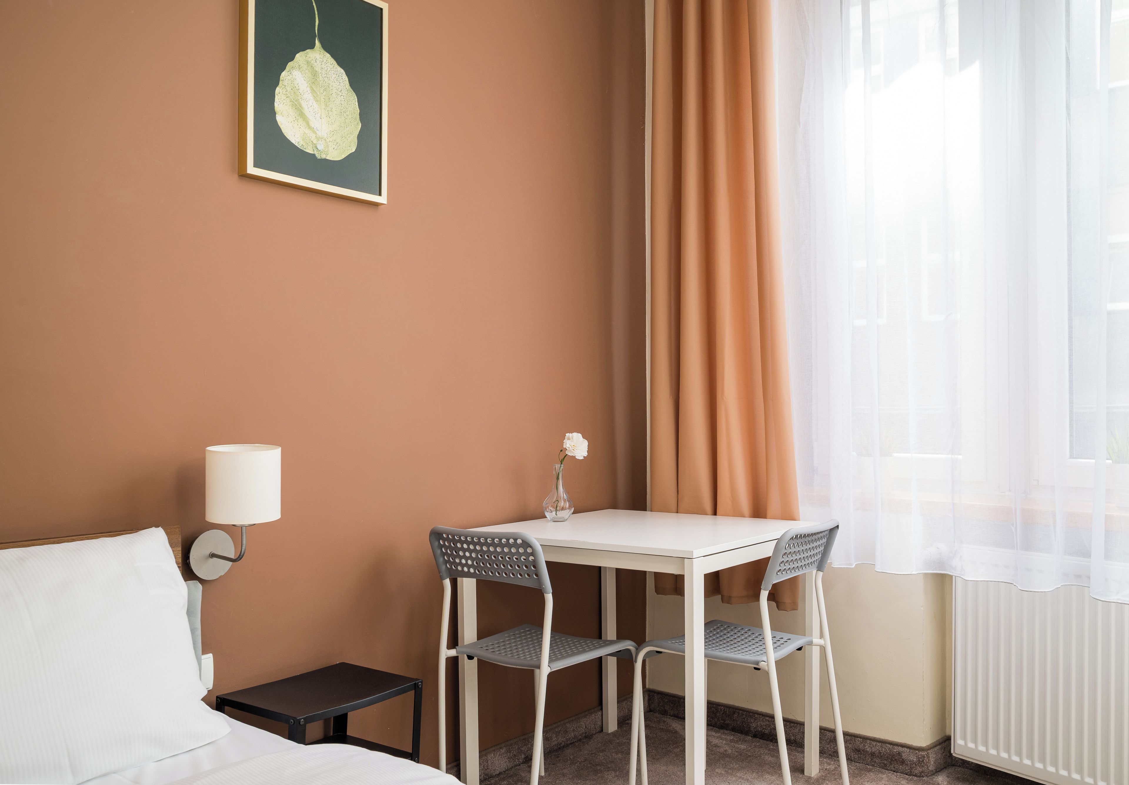 Habitación de hotel con cama, una mesita pequeña con dos sillas, una mesita de noche con lámpara, un cuadro en la pared y una ventana con cortinas translúcidas.