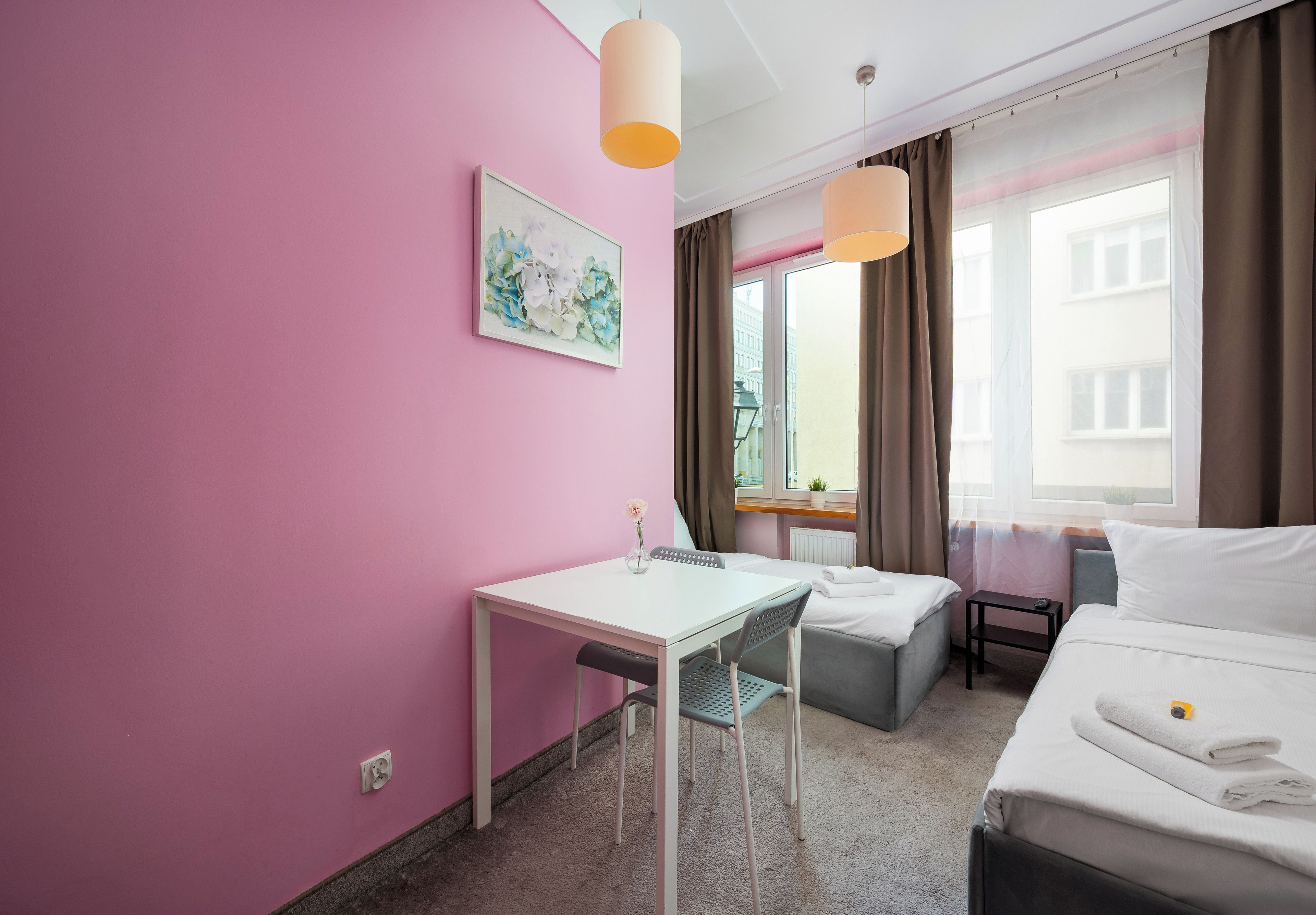 A hotel room with a pink wall, a bed, a desk with a chair, a window with curtains, and a painting on the wall.
