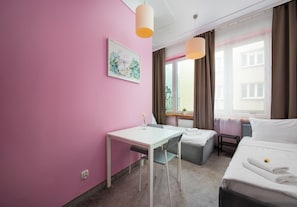 Ett hotellrum med en rosa vägg, en säng, ett skrivbord med en stol, ett fönster med gardiner och en tavla på väggen.