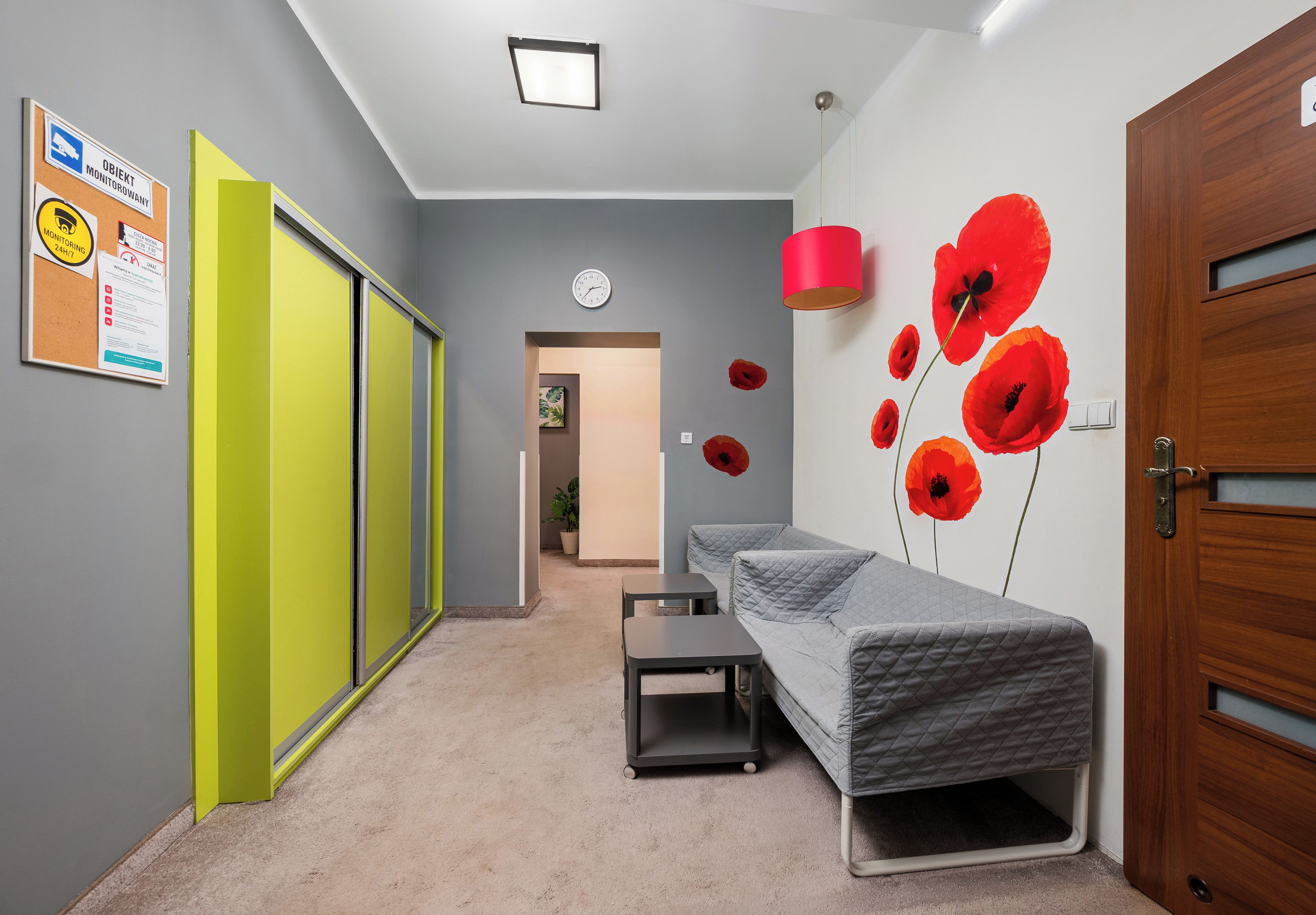 Un pasillo con una puerta verde, un reloj de pared, una cama con cubrecama acolchado y una lámpara roja.