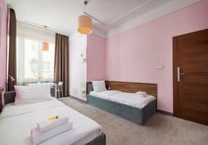 Ein Hotelzimmer mit zwei Betten, einer Holztür, einem Schreibtisch mit Stuhl sowie einem Fenster mit Vorhängen.