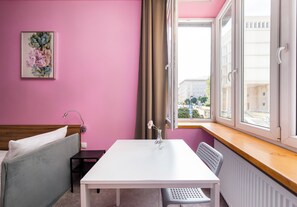 Ein Zimmer mit rosa Wänden, einem Bett mit grauer Bettwäsche, einem weißen Tisch mit einer Vase und einem Fenster mit Blick auf Gebäude.
