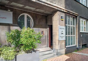 Um edifício com uma placa indicando ser uma pensão. Há uma escadaria que leva à entrada e uma pequena janela com uma placa em cima.