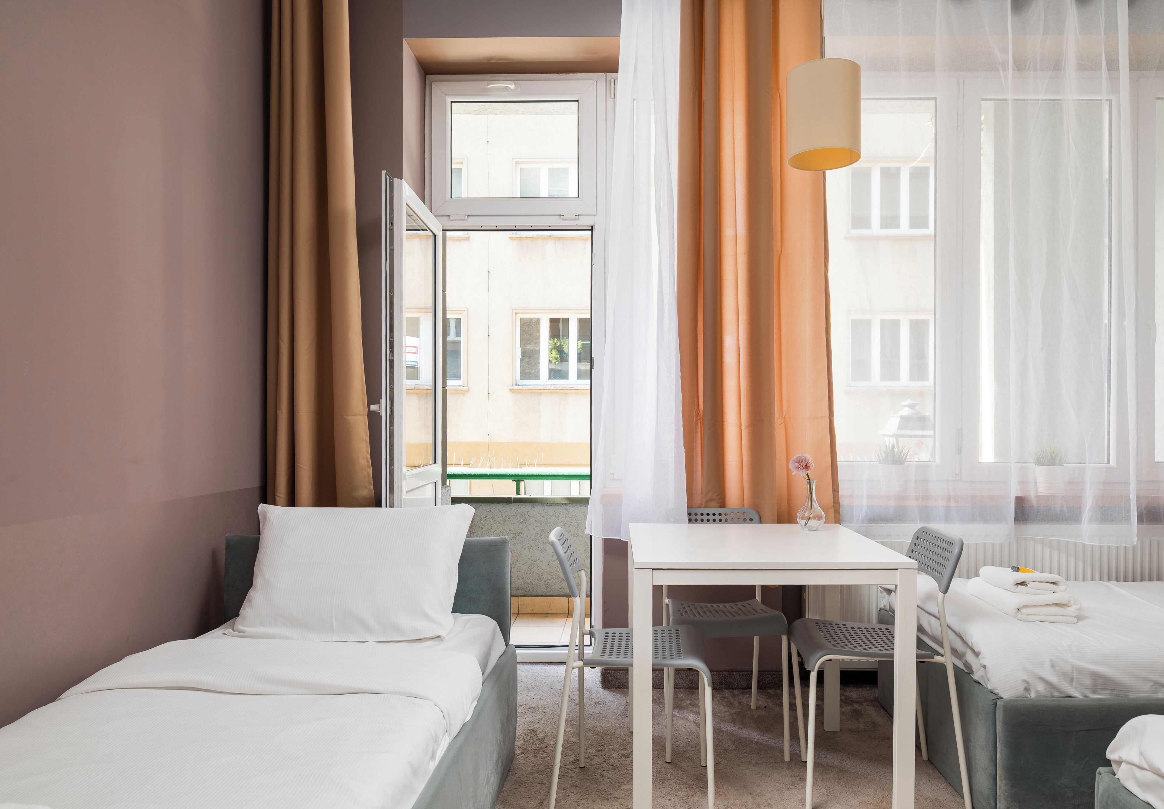 Een hotelkamer met een bed, een bureau en een stoel. Er is een raam met gordijnen, een radiator en een plant op de vensterbank.