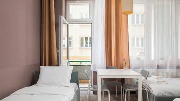 Een hotelkamer met een bed, een bureau en een stoel. Er is een raam met gordijnen, een radiator en een plant op de vensterbank.