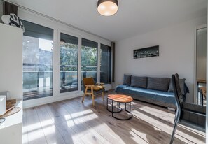 Ein modernes Wohnzimmer mit einer Couch, einem Sessel, einem kleinen Tisch und einem großen Fenster mit Blick auf Bäume und Gebäude.