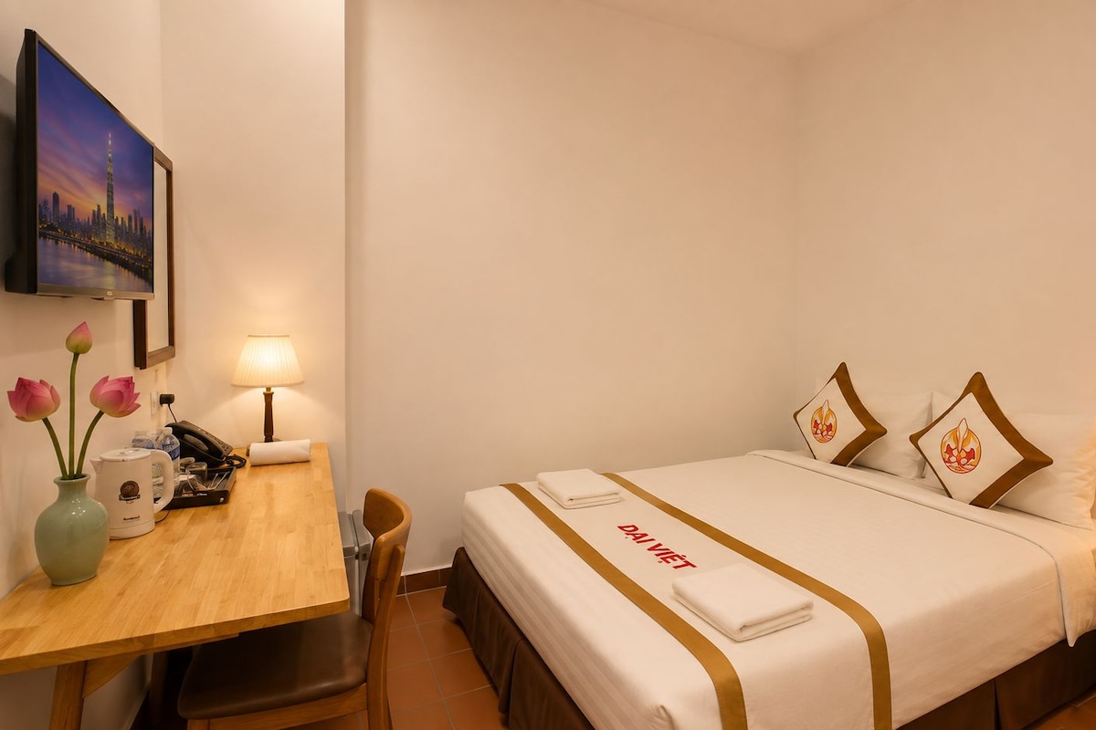 Ein Hotelzimmer mit einem Bett, Nachttischen, einem Schreibtisch, einem Stuhl, einer Blumenvase und einem Bild an der Wand.