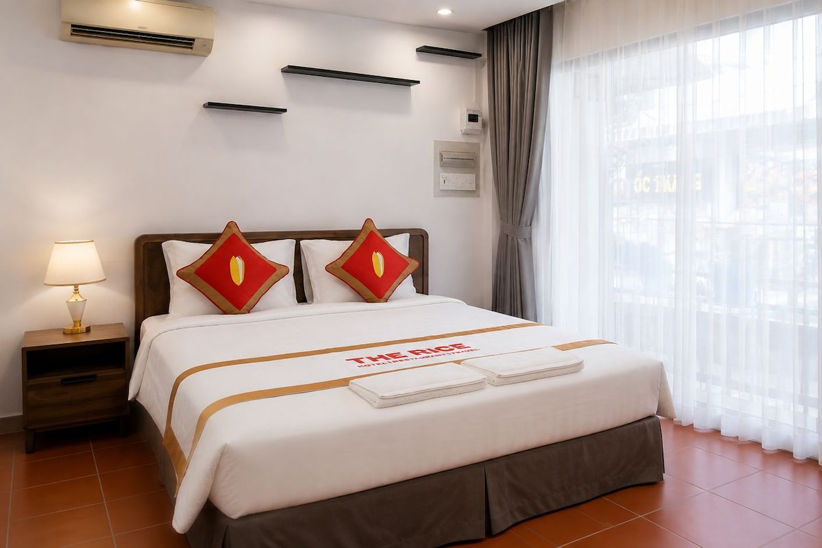 Habitació d'hotel amb llit, taulella, làmpada, finestra amb cortines i aires condicionadors penjats a la paret.