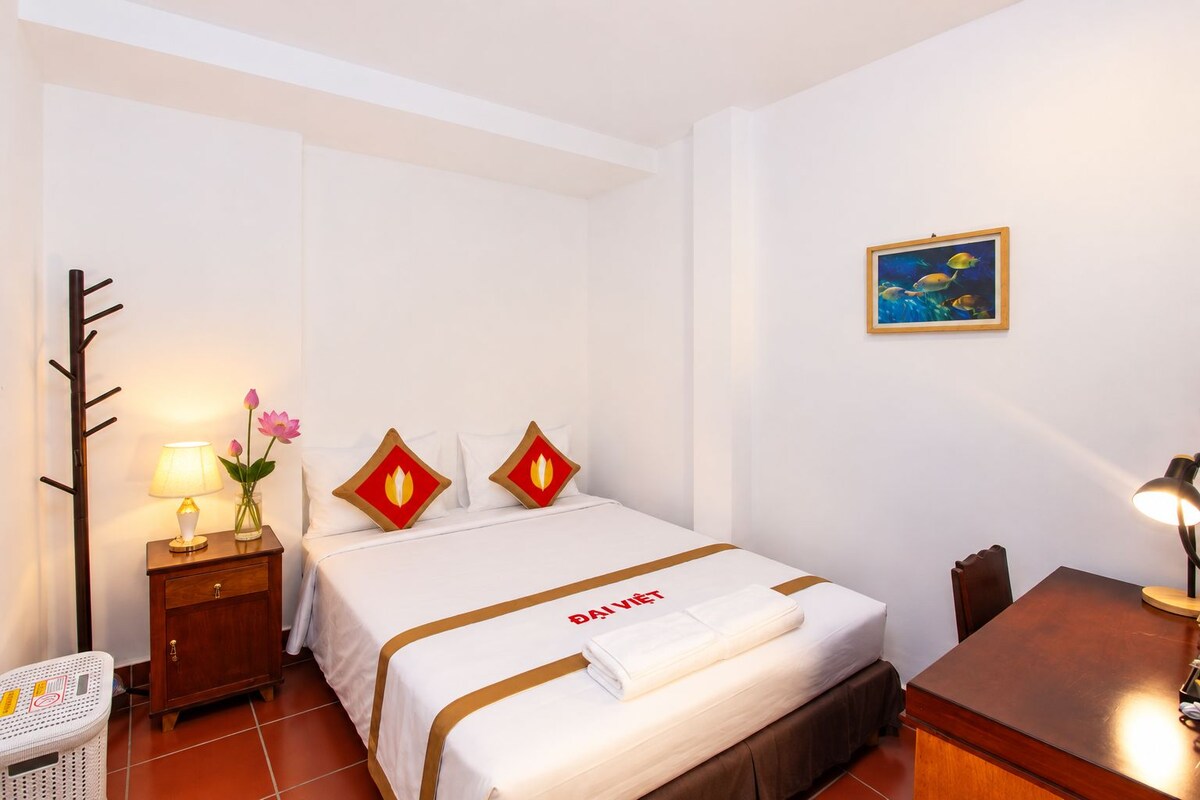 A hotel room with a bed, a nightstand with a lamp, a chair, a desk, and a picture on the wall.