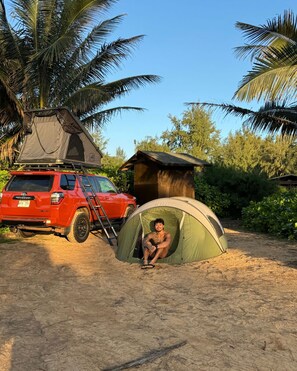 A red SUV parked on a sandy area with a tent and a small wooden structure nearby.