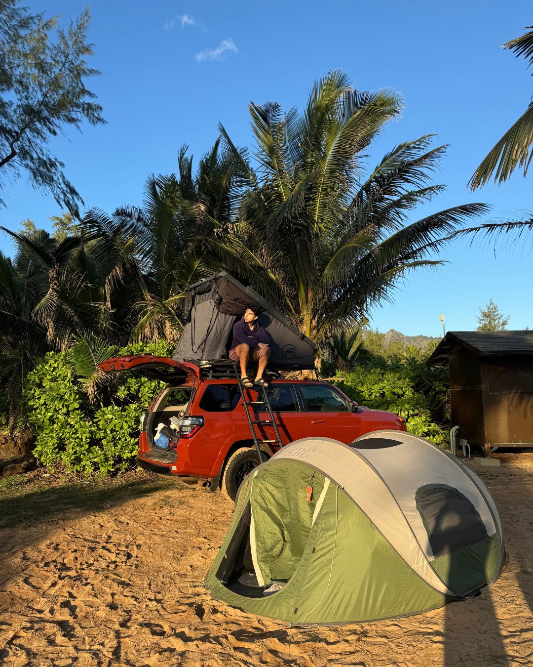 A red SUV parked on sandy ground with a tent and a person standing on its roof.