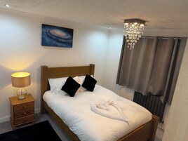 Una cama bien hecha con ropa de cama blanca y almohadas negras, un cabecero de madera, una mesita de noche con una lámpara, un cuadro de una galaxia en la pared y un candelabro.