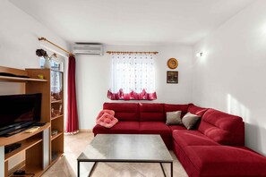 Ein Wohnzimmer mit einem roten Sofa, einem Couchtisch, einem Fernseher auf einem Holzgestell und einem Fenster mit durchsichtigen Vorhängen.
