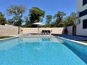 A swimming pool with a patio area, outdoor furniture, and a large tree in the background. - Holiday apartment with shared use of pool (Polje/Krk)