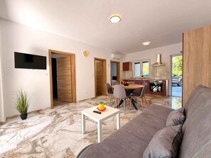 Een moderne woonkamer met een grijze bank, een witte bijzettafeltje en een eethoek met stoelen. Een keuken is zichtbaar door een open deur, en er hangt een televisie aan de muur.