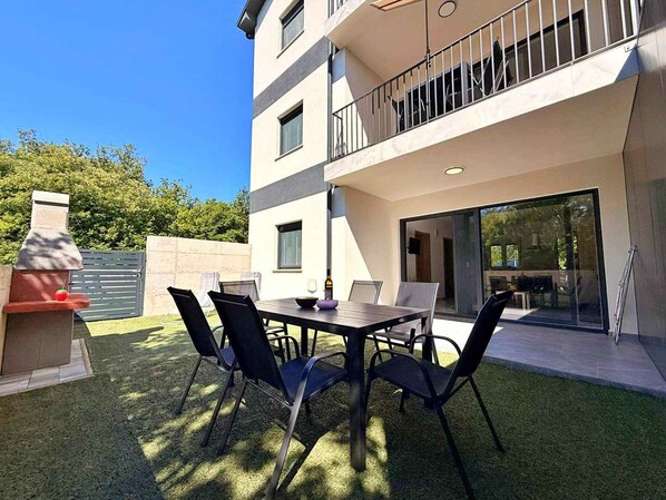 A modern outdoor dining area with a table and chairs, a barbecue grill, and a glass door leading to a balcony. - Holiday apartment with shared use of pool (Polje/Krk)