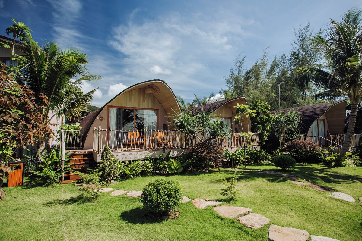 A bungalow-style accommodation with a curved roof, a balcony with a wooden railing, and a garden with grass and rocks.
