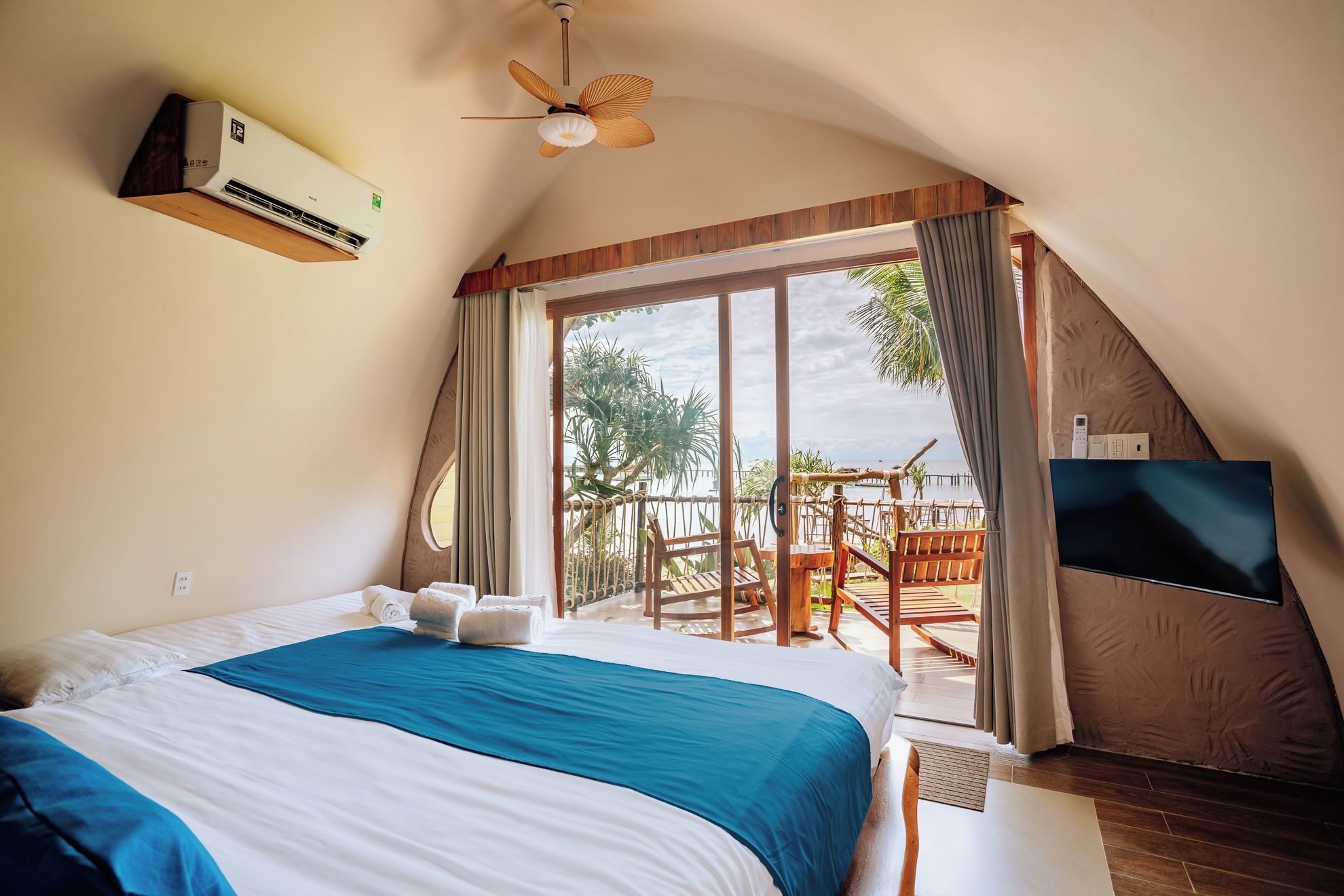 Un dormitorio con una cama grande, ventilador de techo, unidad de aire acondicionado, televisor y balcón con vista al mar.
