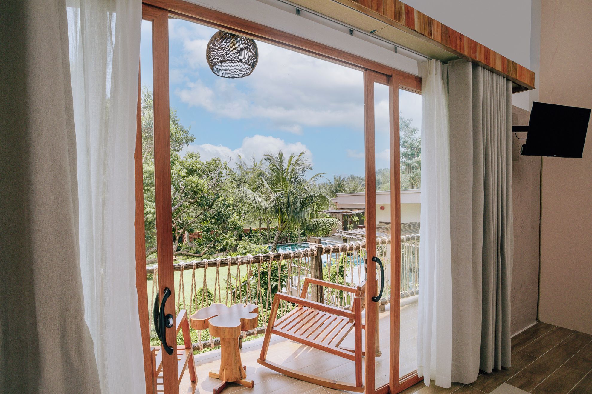 A balcony with a wooden swing chair, a small table, and a view of a pool and greenery.