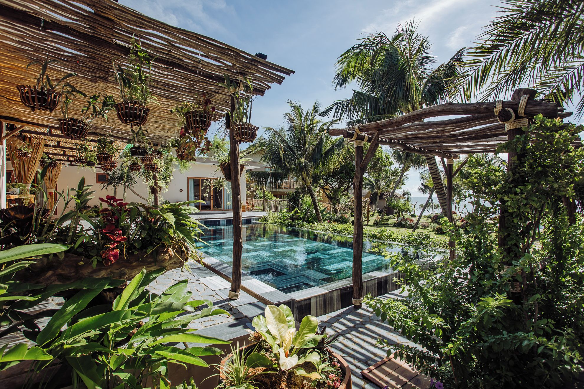 Un área de piscina con una pérgola de madera, exuberantes plantas tropicales y vistas despejadas al océano.