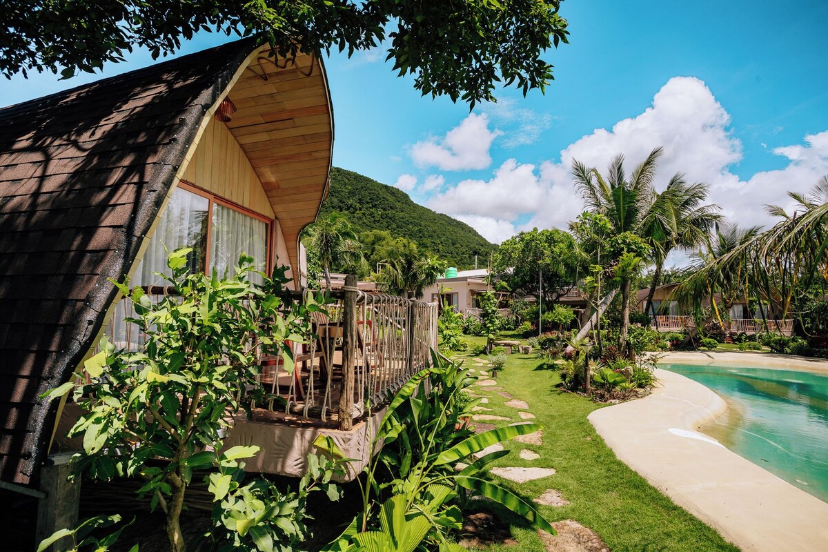A wooden house with a balcony, surrounded by lush greenery and a pool.