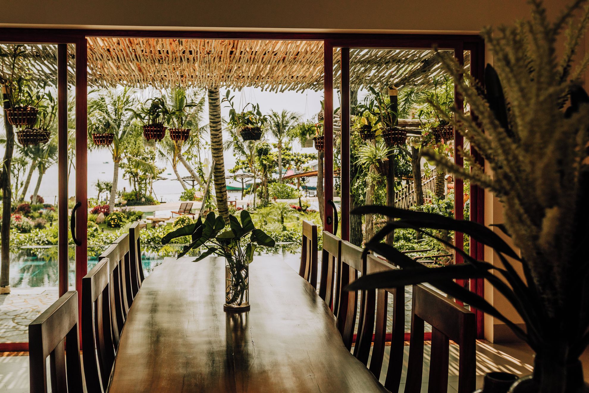 Una mesa de comedor de madera con sillas, una planta colgante y vistas a un jardín tropical con palmeras.