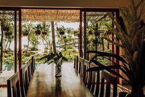 A wooden dining table with chairs, a hanging plant, and a view of a tropical garden with palm trees.