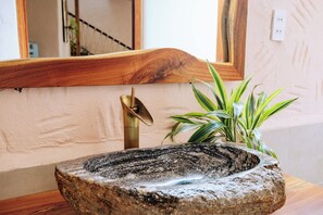 Uma bacia de pedra com torneira de latão, bancada de madeira e planta em vaso com folhas listradas.