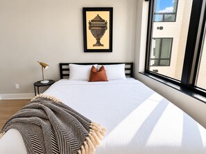 A large bed with white bedding, a framed artwork above it, a side table with a lamp, and a view of a building through the window. - Sleepover | Dashing Studio + Gym/Sauna + Omaha (Omaha)