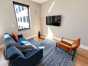 A modern living room with a sofa, a coffee table, a TV mounted on the wall, and a window with a view of a building. - Sleepover | Dashing Studio + Gym/Sauna + Omaha (Omaha)