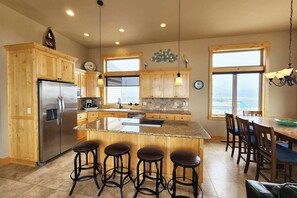 A kitchen with wooden cabinets, a stainless steel refrigerator, a large island with stools, and a dining area with chairs. - Luxurious Home Sits Right on the Beach! Pet Friendly! (Waldport)