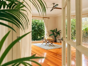 A room with a wooden floor, a ceiling fan, a table with a cushion, and a view of greenery outside. - Warrandyte Bush Retreat  6BR with Private Pool (Warrandyte)