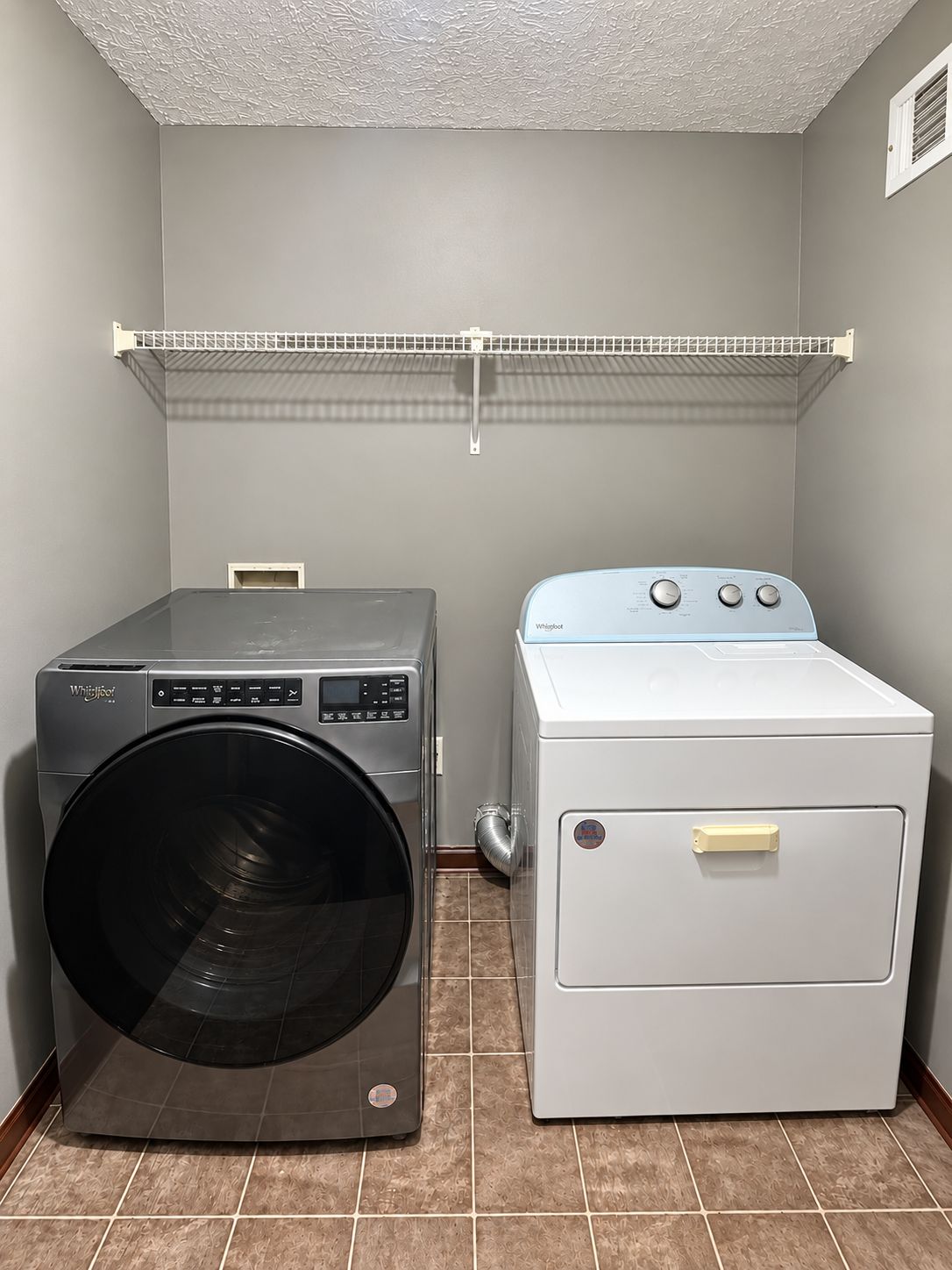 A laundry room with a front-loading washer and a top-loading dryer, a metal shelf above, and a vent on the wall.
