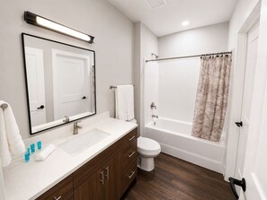 A modern bathroom with a white sink, mirror, and toilet, a wooden vanity, and a shower with a curtain. - Sleepover | Peaceful Studio + Gym/Sauna - Omaha (Omaha)