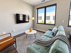 A living room with a sofa, a coffee table, a TV mounted on the wall, and a window with a view of a building outside. - Sleepover | Peaceful Studio + Gym/Sauna - Omaha (Omaha)
