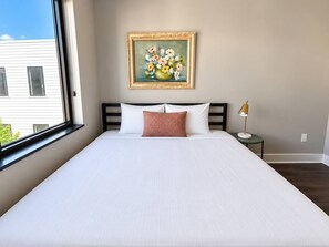 A hotel room with a large bed, a painting of flowers on the wall, a window, and a bedside lamp. - Sleepover | Peaceful Studio + Gym/Sauna - Omaha (Omaha)