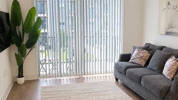 A modern living room with a grey sofa, a large plant, a ceiling fan, and a view through floor-to-ceiling windows.