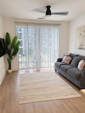 En moderne stue med en grå sofa, en stor plante, en loftsvifte og udsigt gennem gulv til loft-vinduer.