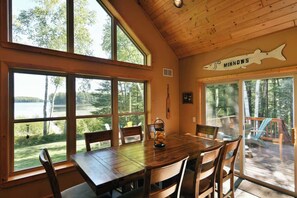 A wooden dining room with large windows overlooking a lake, a fish-shaped sign on the wall, and a view of the deck. - Musky Haven-Your Northwoods Gathering Spot (Hayward)