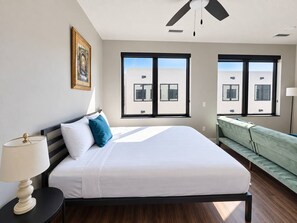 Ein modernes Hotelzimmer mit einem großen Bett, einem Nachttisch, einer Lampe, einem Fenster mit Blick auf Gebäude und einem Deckenventilator.