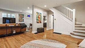 A spacious living room with a wooden floor, a brown leather sofa, a patterned rug, and a staircase with white railings. - This NEW "Old" House• Broad Ripple Home Steps From Dining, Nightlife & The Monon (Indianapolis)