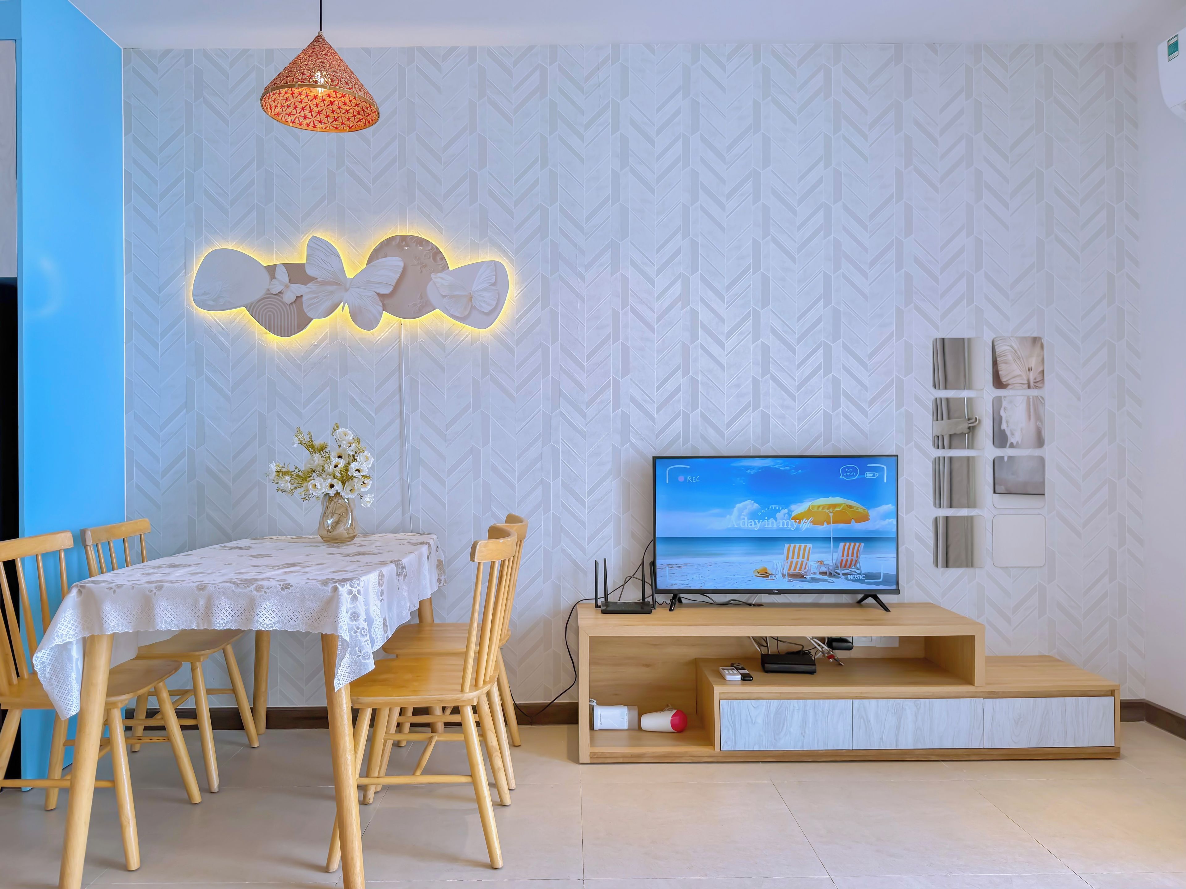 Un área de comedor con una mesa y sillas, un televisor sobre un soporte de madera, y una pared decorativa con un diseño geométrico.