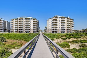 A wooden boardwalk leading to a beachfront hotel with multiple balconies and a clear blue sky. - Oceanfront | Private Balcony | Luxury Ocean Place Condo Next to the Ritz (Fernandina Beach)