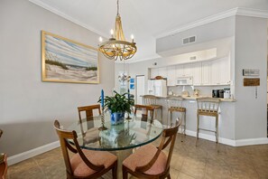 A dining area with a glass table, wooden chairs, a chandelier, and a painting on the wall. - Oceanfront | Private Balcony | Luxury Ocean Place Condo Next to the Ritz (Fernandina Beach)