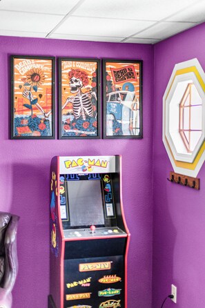 Ein Zimmer mit einem Pac-Man-Spielautomaten, gerahmten Postern an der Wand und einer violetten Wand mit Struktur.