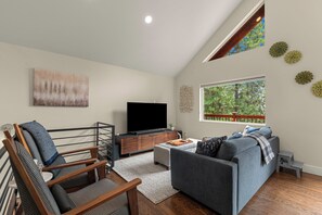 A modern living room with a flat-screen TV, a grey sofa, a wooden chair, and a small table. - Turkey Trot Lodge | Private Hot Tub, 2 Miles to Downtown Leavenworth (Leavenworth)