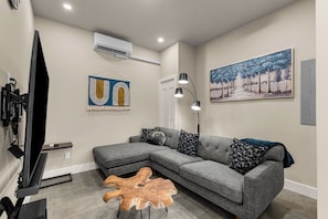 A modern living room with a sectional sofa, a wooden coffee table, a painting, and an air conditioning unit. - Turkey Trot Lodge | Private Hot Tub, 2 Miles to Downtown Leavenworth (Leavenworth)