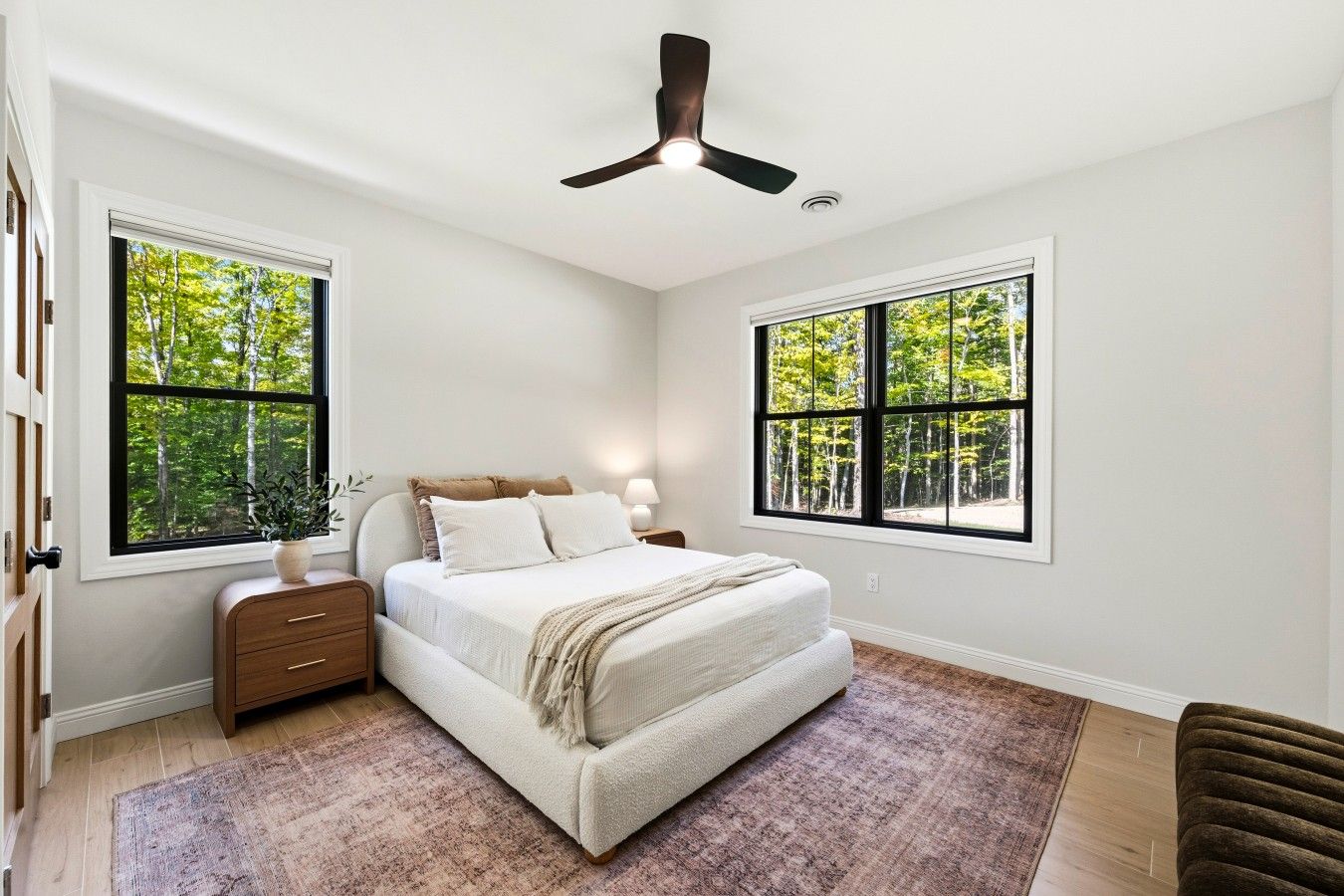 A bedroom with a bed, nightstands, a ceiling fan, and large windows with a view of trees.