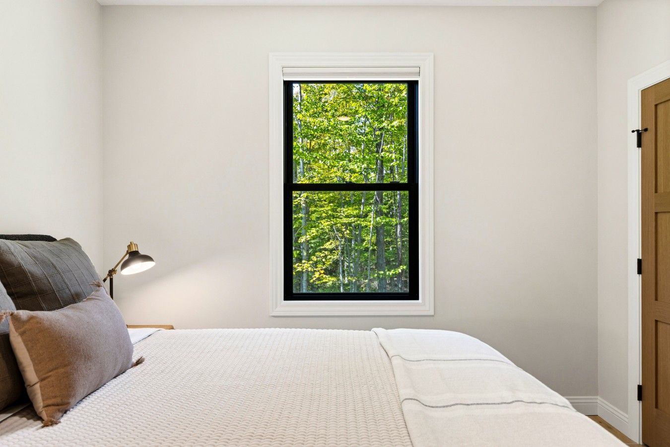 A bedroom with a bed, pillows, a nightstand, a lamp, and a window with a view of trees.