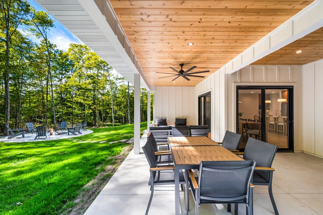 An outdoor patio with a long wooden table and chairs, surrounded by a lush green lawn and trees.